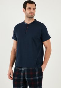 Cămașă albastră cu mânecă scurtă, cu guler butonat și detalii în carouri la guler, asociată cu pantaloni de pijama în carouri, în nuanțe de albastru, roșu și alb.