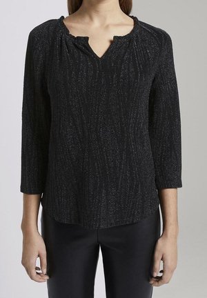 Žena nosící černý texturovaný top s dlouhým rukávem a malým výstřihem ve tvaru V a černé kalhoty, stojící na jednobarevném pozadí.