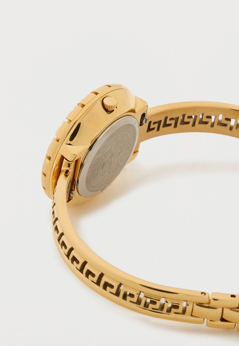 Horloge van goudkleurig metaal met een ronde kast, een gestructureerde kroon, en een patroonriem met uitsparingen langs het bandje.