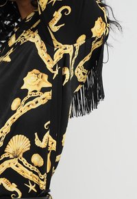 Svart tyg med gula snäckor, sjöstjärnor och kedjemönster, med långa svarta fransar på ärmarna.