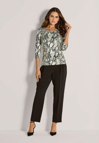 Frau in einem schlangenbedruckten Oberteil mit Dreiviertelärmeln, schwarzen, taillierten Hosen und schwarzen Ankle-Strap-Heels, die vor einer hellen Wand steht.