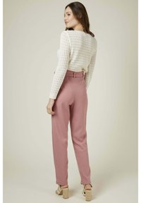 Pull en tricot blanc à manches longues, associé à un pantalon rose taille haute. La mannequin se tient devant un fond neutre, montrant le tombé complet du vêtement.