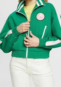 Grön zip-up jacka med åtsittande midja, utrustad med vita och rosa randdetaljer, en rund patch och ribbade ärmslut. Bärs över en gul topp.