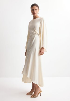 Kobieta stojąca w kościelnobiałej asymetrycznej sukience z długimi rękawami i marszczeniami, nosząca złote kolczyki koła i sandały na obcasie.