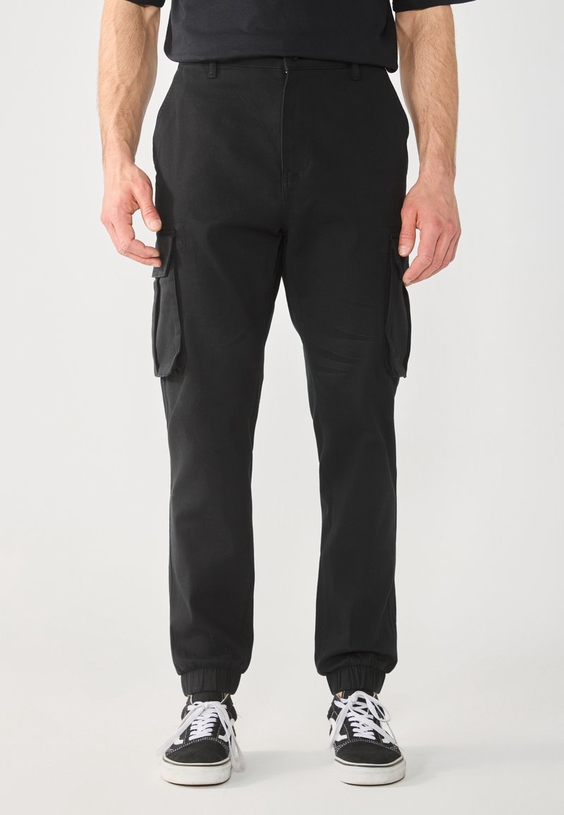 Homme portant un pantalon cargo noir avec poches latérales et des baskets noir et blanc, debout sur un fond clair uni.