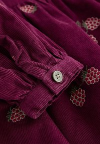 Bordový kordový materiál s textúrovanými prúžkami, so zapínaním na gombík a vyšívanými malinovými vzormi v červenej a zelenej farbe.