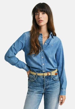 Donna con lunghi capelli castani che indossa una camicia di denim abbottonata, cintura chiara intrecciata e jeans blu sbiaditi, in piedi con una mano sul fianco.