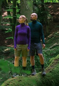 Kaksi henkilöä seisoo sammalen peittämällä puunrungolla metsässä. Toinen on pukeutunut purppuraväriseen pitkähihaiseen paitaan ja vaaleisiin shortseihin; toinen pitää päällään tummanvihreää paitaa ja shortseja.