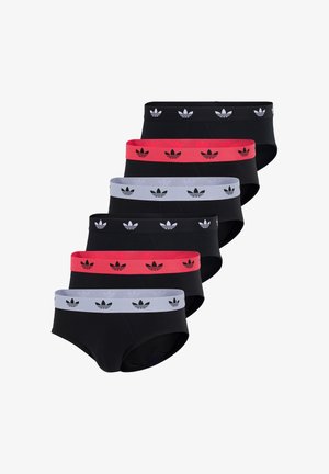 Młody mężczyzna z piegowatą skórą i warkoczami, ubrany w czarne bokserki z białym paskiem w logo Adidas, stoi boso na białym tle.