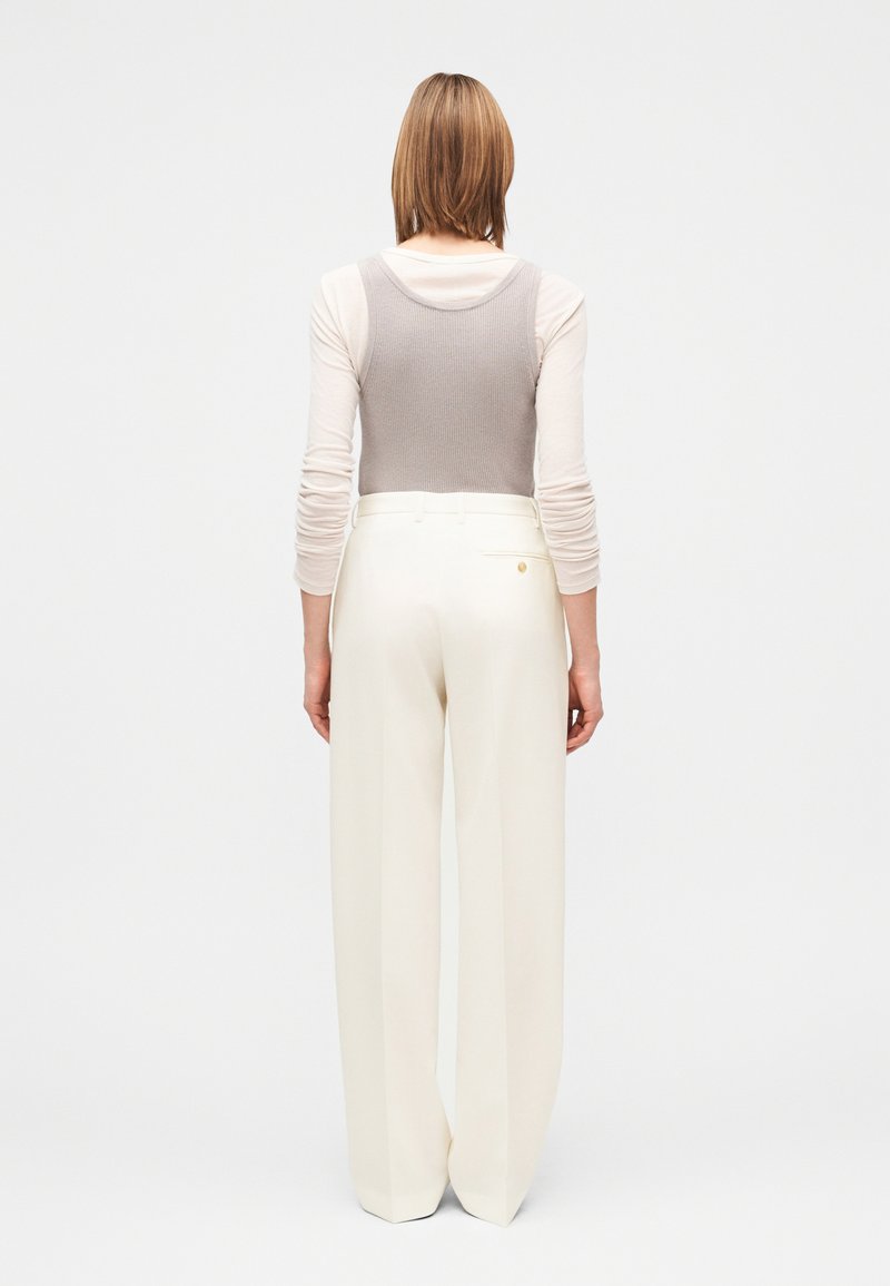 Débardeur gris clair côtelé superposé à une chemise crème à manches longues, associé à un pantalon blanc taille haute à jambes larges avec une poche arrière.