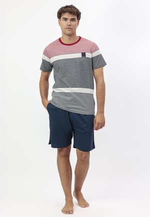 Joven de pie descalzo, vistiendo una camiseta de bloques de colores con pantalones cortos azul marino, una mano en el bolsillo, fondo neutro.