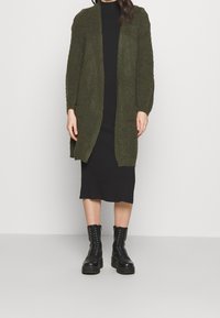 Olivgrön stickad kofta, öppet fram, texturerade ärmar, bärs över en svart ribbad klänning, kombinerad med svarta snörda kängor.