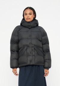 Person iført en sort dunjakke med høj krave og store frontlommer, stående mod en ensfarvet hvid baggrund.