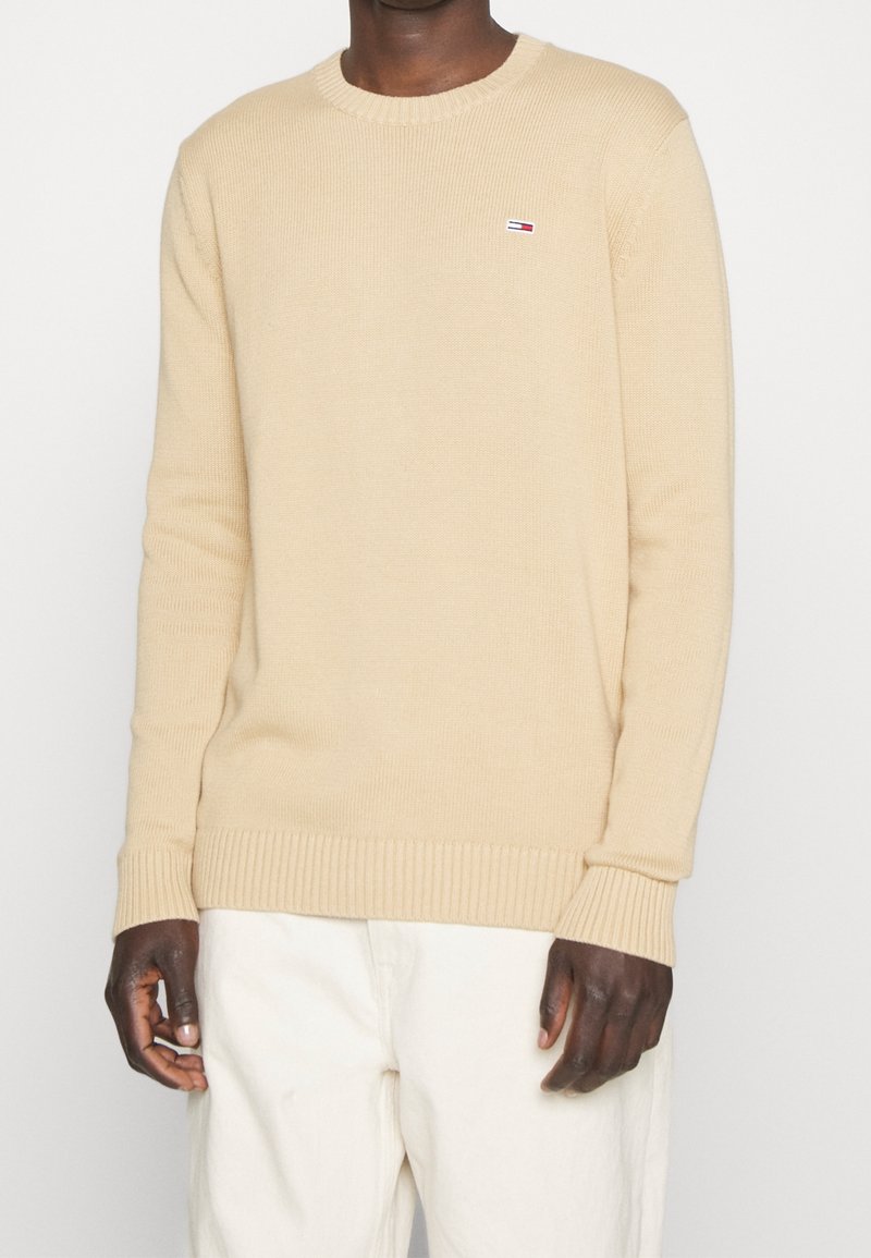 Pull en tricot beige avec un col ras du cou et des poignets côtelés. Présente une petite étiquette logo sur la poitrine. Porté avec un pantalon de couleur claire.