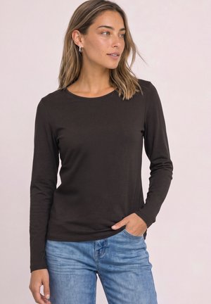 Femme aux cheveux mi-longs portant des boucles d'oreilles anneaux en argent, une chemise noire à manches longues et un jean bleu clair, une main dans la poche.