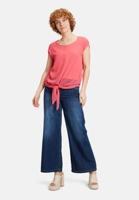 Top rosa a maniche corte con nodo frontale, abbinato a jeans in denim scuri a gamba larga e scarpe con tacco platform di colore chiaro. Tessuto morbido, vestibilità rilassata.