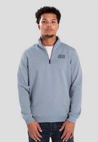 Ljusblå kvartdragning med mjuk textur och ribbad mudd. Har en svart logotyp på vänster bröst. Bärs över en vit T-shirt.