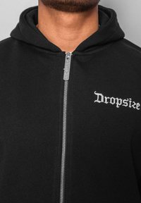 Sweat à zip noir fabriqué en tissu doux, avec une fermeture éclair argentée et "Dropsize" brodé en blanc sur le côté gauche de la poitrine.