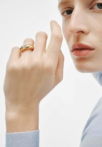 Anillo de oro con un diseño suave y redondeado, que presenta una piedra clara en la parte superior, llevado en una mano de tono de piel claro. Manga azul suave visible.