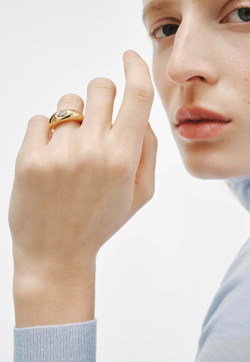 Anillo de oro con un diseño suave y redondeado, que presenta una piedra clara en la parte superior, llevado en una mano de tono de piel claro. Manga azul suave visible.