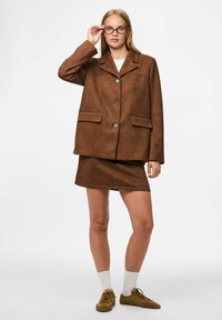 Smeđa zamšna jakna s izrezanim ovratnikom, prednjim gumbima i džepovima, uparena s odgovarajućom smeđom suknjom i svijetlim cipelama. Bijele čarape su vidljive.