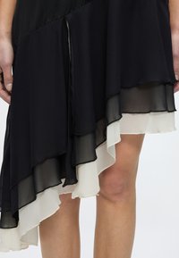 Μαύρη και λευκή στρωματοποιημένη ασύμμετρη φούστα που φοριέται από άτομο, με εμφανή χέρια και εκτεθειμένα γόνατα σε ουδέτερο φόντο.