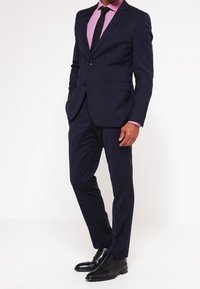 Costume bleu marine à coupe ajustée, avec une fermeture à deux boutons, une chemise rose et des chaussures habillées noires. Texture de tissu lisse.