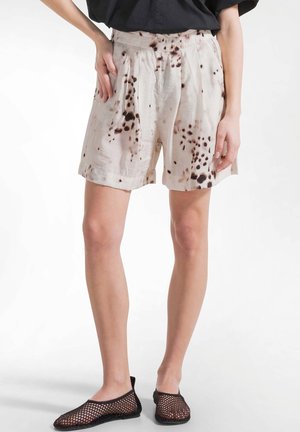 Femme portant un short ample beige taille haute avec des taches brun foncé, associé à des chaussures slip-on en maille noire et un haut noir.