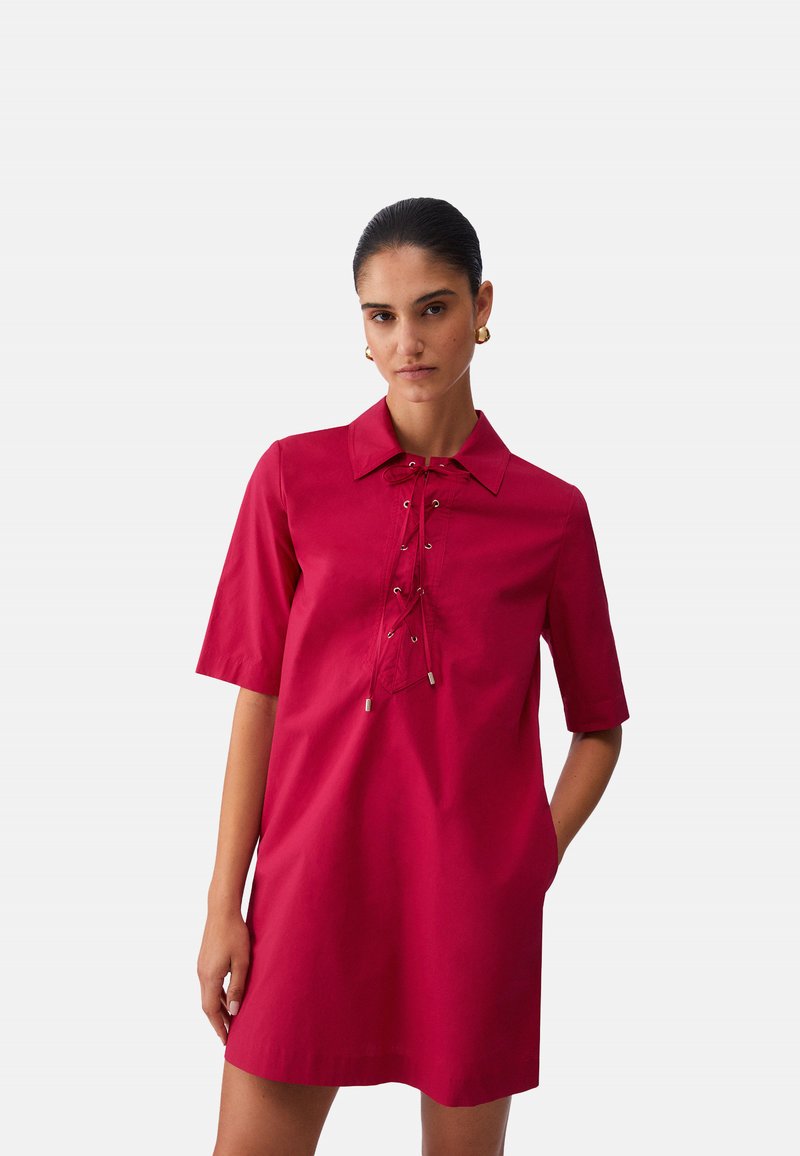 Femme portant une robe rouge à manches courtes avec un détail à lacets sur le devant et un col, debout avec une main dans la poche contre un arrière-plan uni.