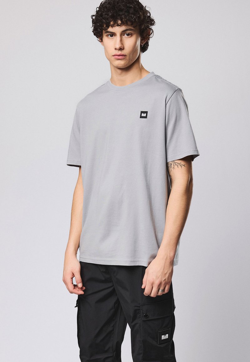 Jeune homme aux cheveux bouclés portant un t-shirt gris clair et un pantalon cargo noir, debout devant un fond clair uni.