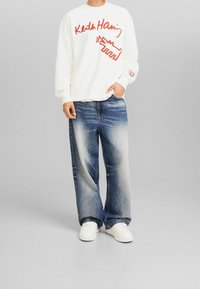 Vit sweatshirt med röd grafisk text och mönster. Bärs med ljusblå vida jeans. Vita sneakers fulländar outfiten.