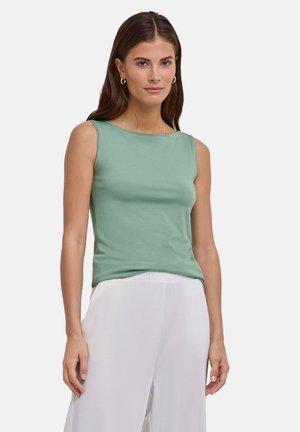 Donna con capelli lunghi castani, che indossa una canotta verde e pantaloni bianchi, in piedi davanti a uno sfondo chiaro e uniforme.