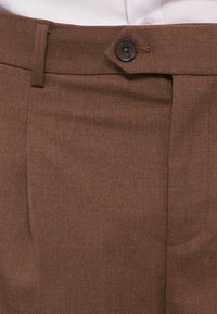 Braune Hosen aus strukturiertem Stoff, mit einer Vordertasche, Ein-Knopf-Verschluss und Faltenverlauf entlang der Beine.