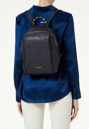 Sac à dos en cuir noir texturé avec une forme arrondie, présentant des accents en métal doré et deux bretelles réglables. Compartiments zippés visibles.
