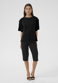 Haut noir à manches courtes avec un col rond et des détails de manches repliées, associé à un pantalon noir longueur genou et des sandales noires à bout ouvert.