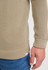 Beige geribbelde sweater met een gestructureerd oppervlak, strakke boorden en een label aan de onderrand. Gecombineerd met blauwe jeans, wat een casual silhouet toont.
