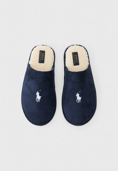 Tumši zili zamšāda čības ar mīkstu krēmbaltu mākslīgās kažokādas oderi un baltu izšūtu polo logotipu priekšpusē. Atvērta aizmugure, viegli uzvelkamas.