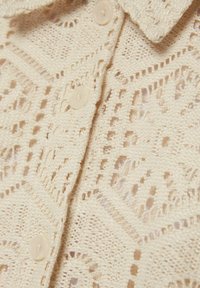 Blusa de encaje en suave beige con un patrón geométrico, que presenta un cuello y botones redondos, mostrando una textura intrincada y un diseño de punto abierto.