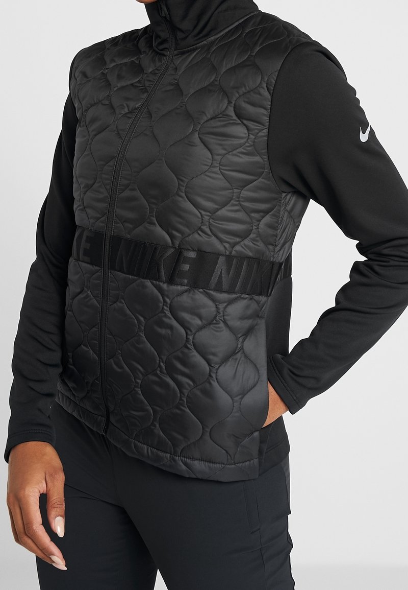 Personne portant un gilet matelassé en nylon noir avec le logo "NIKE" sur un t-shirt noir à manches longues avec un petit swoosh blanc Nike sur la manche.
