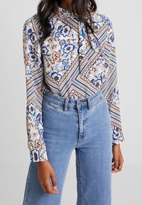 Halenka s dlouhým rukávem z lehké látky s květinovým a geometrickým vzorem v modré, bílé a hnědé barvě. Kombinováno s modrými džíny s vysokým pasem.