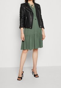 Chaqueta de cuero negra de motociclista sobre un vestido verde hasta la rodilla con falda de capas y un lazo en el escote. Sandalias negras de tacón alto.