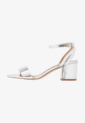 Silver metallic heeled sandal with a wide toe strap, ankle strap, and block heel. Smooth texture with a shiny finish and minimalistic design.