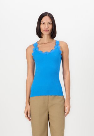 Sinine ribitud tank ülikond, millel on laineline pitsääre kaelaavardil ja õlgadel, omab kätšitud disaini ja sirget äärt. Kombineeritud beežide pükstega.