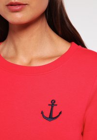 Červená mikina s kulatým výstřihem, zdobená námořnicky modrým vyšívaným kotvou na levé straně hrudi. Vyrobená z měkké, texturované látky.