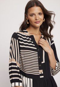 Sukienka w czarno-beżowe paski z zapięciem na guziki z przodu, bez kołnierzyka i rękawami o długości trzech kwart. Posiada plisowany dół oraz różnorodne wzory pasków.