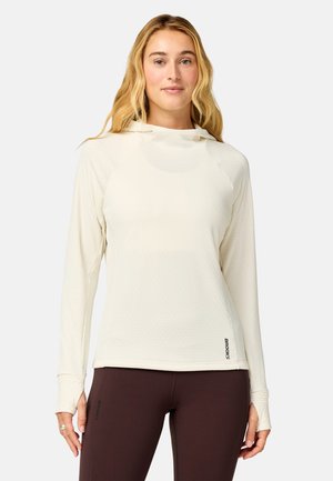 Kvinde med langt blondt hår iført en cremefarvet hættetrøje med lange ærmer og mørkebrune leggings, stående mod en ensfarvet baggrund.