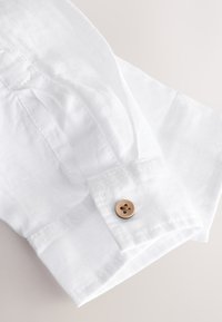 Manche de chemise en lin blanc avec un seul bouton beige sur le poignet, présenté sur un fond clair et neutre.