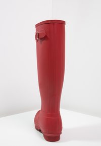 Botte de pluie en caoutchouc rouge avec une finition lisse, design montante jusqu'au genou, et accent avec boucle sur le côté. Dispose d'une semelle texturée pour l'adhérence.