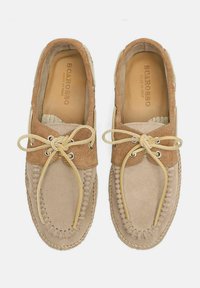 Mocasines de ante beige con un acento texturizado marrón, cordones beige y costuras decorativas a lo largo de los bordes. El interior presenta una marca en relieve.