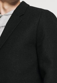 Černé kostkované sako z texturované směsi vlny s klopovým límcem a střih přiléhavý, vrstvené přes šedou mikinu s kapucí.